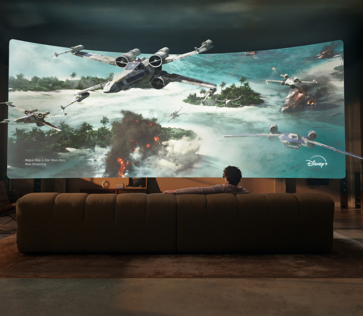 Une personne assise sur un canapé et portant des lunettes Meta VR fait face à un grand écran virtuel incurvé montrant une bataille de X-wing de Rogue One sur Disney+