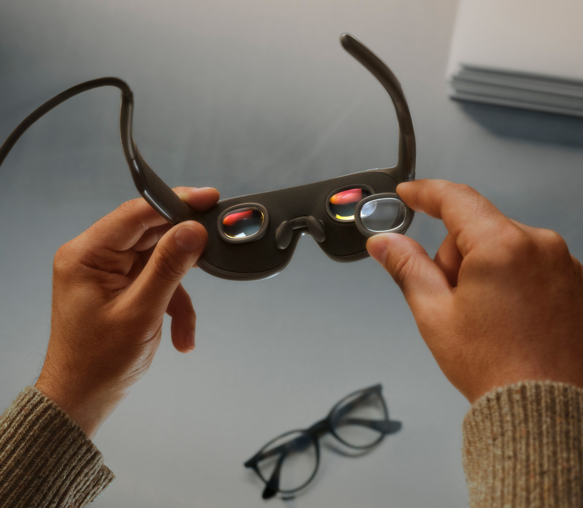 Deux mains tiennent les lunettes Meta VR à l'envers, en plaçant un insert de prescription ovale transparent sur l'une des deux petites lentilles.